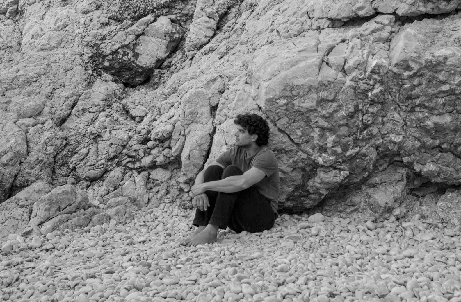 Stephan standing simply on a pebble beach in Marseille, a large limestone cliff rising behind him.